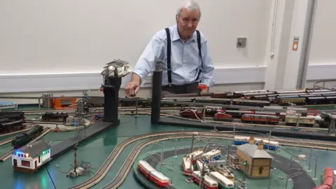 Rod Moore Rod Moore with his layout which is large and has winding tracks with houses dotted around. Rod is wearing a blue shirt with black braces and has a sheepish expression on his face as he presses a button to operate the model. 