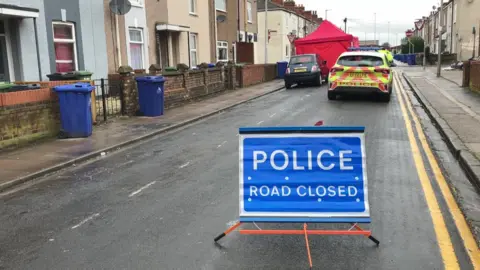 A blue sign reading "POLICE ROAD CLOSED" in white writing and capital letters. It is in the middle of the road. A marked police car and a red tent can be seen in the distance. Residential houses line the street. 