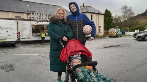 A woman with blonde hair wears a long dark green puffa jacket. She stands next to a teenage boy who is taller than her and wears a black and blue padded jacket and holds a football. The woman holds the handle of a red pram in front of her with a young child in it. They stand in a car park on a grey day in front of a food van.