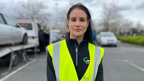 A woman in high-vis tabard with long dark hair with a seized vehicle on a low loader on the right hand side of the picture.