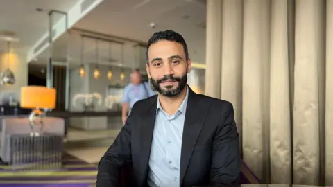 Mazen Fadhl wears a black suit and light blue shirt is seated at a table in a modern, well-lit room with purple and yellow carpet. In the background, there is another person standing near a lounge area with contemporary furniture and hanging lights.