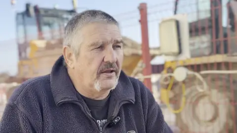 Alabaré A man with grey hair and a moustache, looking off the camera. He is wearing a black fleece. 