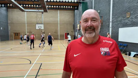 Bob Mitchell wearing a red Nike t-shirt while standing in a sports hall. He is bald with a stubbly white beard. 