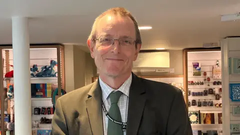 Jack Maclean/BBC Dr Robin Hanley standing inside a room in Norwich Castle. He is wearing a dark green suit jacket, with a checked shirt and dark green tie. He is wearing glasses and is looking directly at the camera and smiling.