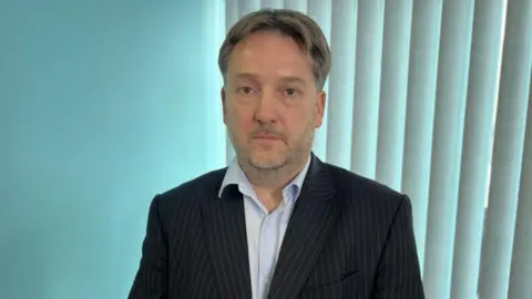 BBC / Fiona Irving A man with grey hair, wearing a black suit and white shirt. He is standing inside an office.