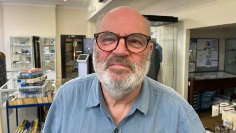A bald man with thin grey hair on the side of his head. He has a grey beard and moustache and is wearing brown glasses. He is wearing a blue denim-type shirt. Behind him are items on display at the museum.