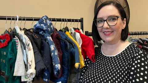 BBC Karen Brown is standing in front of a rack of baby and toddler clothes. She wears black glasses and has bobbed hair. There's a clock on the wall behind her.