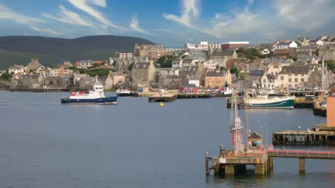 Getty Images Wide view of a coastal harbour with several boats on calm water, including a ferry and fishing vessels, alongside a town of stone buildings on a gentle hillside under a blue sky with scattered clouds.