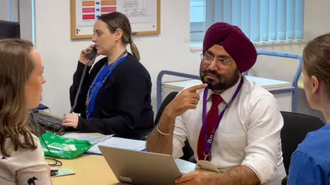 Dr Inder Singh sitting at a desk with a laptop . Beside him is a woman nurse speaking on the phone and across from him are two women listening. Inder is wearing a white shirt and red tie. He has a short beard and is wearing an maroon turban. 