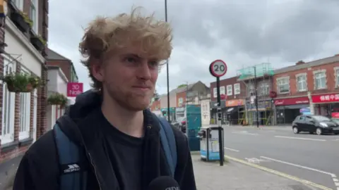A man with blonde hair stood next to a main road. He is wearing a black hoodie and a blue backpack