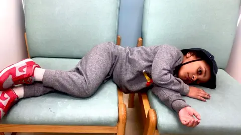 Sinead Picture of Aziyah laying across two green hospital chairs. He is wearing a grey tracksuit and red trainers, and a black cotton hat. He is looking into the distance.