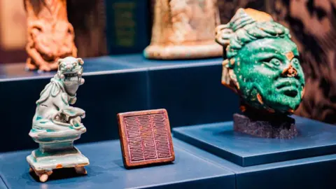 Royal Armouries/Nomad Exhibitions Ceramics in an exhibition about the Mongolians which include a green head statue and a blue figure of an animal. There is a red inscribed square between the two. 