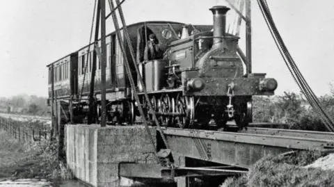 A historic black-and-white photograph showing a steam train crossing a lift bridge on the Hundred of Manhood and Selsey Tramway. The bridge spans a waterway while the locomotive and carriage travel across it.