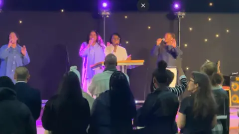 Vigil attendees gathered in a church, facing a stage where four individuals are singing into microphones. The singers are illuminated by purple stage lights. Attendees are standing and appear engaged in the service, some raising their hands.