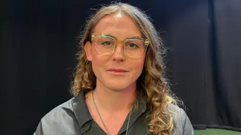 Kate Lankester looking directly down the camera. She is wearing a grey, open-necked, collared top, with a silver chain around her neck, though the pendant is not in the frame. She is wearing square-framed glasses with transparent plastic frames. She has curly, light brown hair that is falling below one of her shoulders. She is sat against a black backdrop.