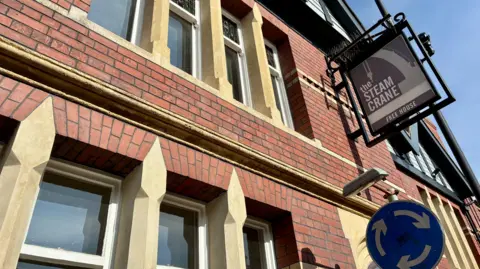An exterior view of the pub, showing the sign for the Steam Crane. It is a redbrick building with white stone window columns.
