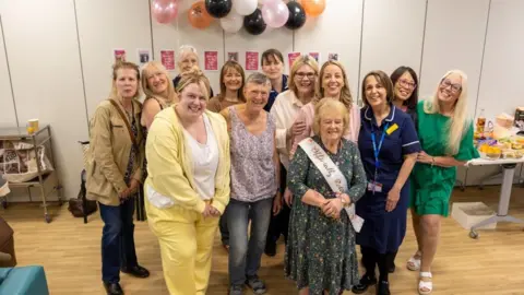 NHS Humber Health Partnership Janet Harley, with short, blonde hair, wearing a green, floral dress and a pink and gold sash. She is surrounded by a group of women. Black, orange, pink and white balloons are stuck to the cream-coloured wall behind her and there is a table of food in the background.