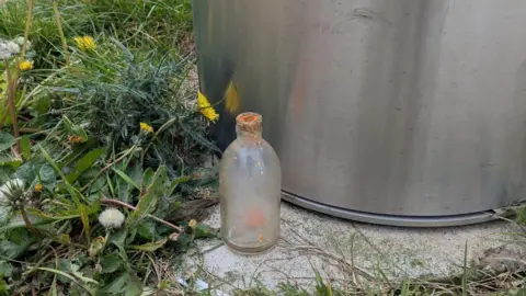 Marie Lovell A glass bottle on concrete next to a metallic bin. The glass is opaque and seems to have orange liquid in it. The top of the bottle is crusted with orange paste.