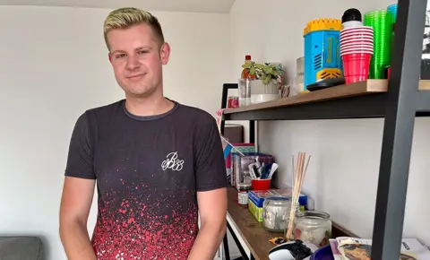 Guy Benson stands successful  his shared ownership level  astatine  Landmark Pinnacle, he's wearing a achromatic  and reddish  T garment  and smiling with blonde hair, adjacent  to a partition  and a acceptable   of shelves with assorted  homely possessions connected  them.