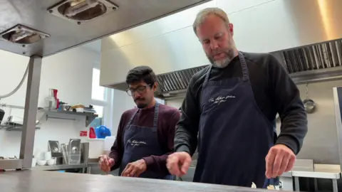 Two men in a white kitchen with silver equipment. They are looking down at a worktop, which is not in frame, and wearing dark blue aprons.