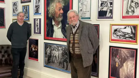 MNH Curator Matthew Richardson, a man with grey hair, on the left and Major Shane Lucas, a man with long grey hair and a beard, on the right stand in front of a wall of paintings.