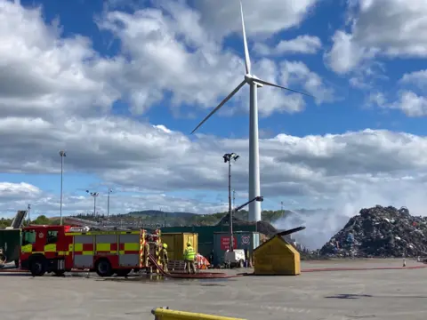 A fire engine is spraying water on a pile of waste that is smoking