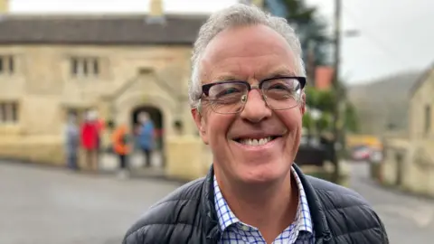 Simon Coombe beaming at the camera in front of the Hop Pole - a yellowed stone building with small vertical windows in clusters of three.