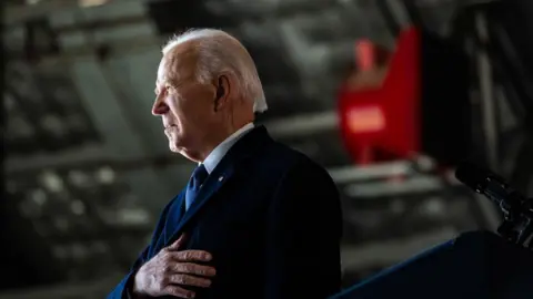 Former US President Joe Biden's side profile. His hand is laid on his chest and he has a dark coat on.