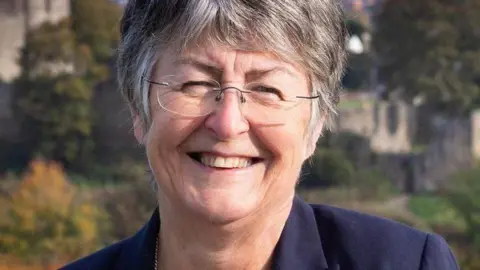 Photo of Heather Kidd smiling in the centre of the screen. She is wearing a navy blue jacket.  