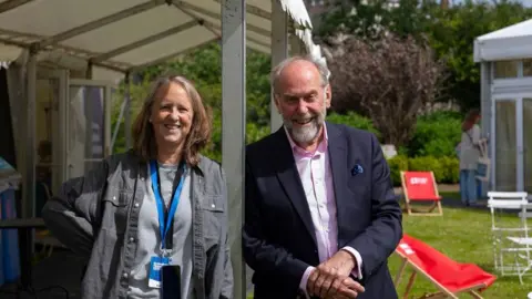 Lloyd Smith Photography Two people stand side by side leaning on a metal marquee pole. A woman on the left has long brown hair and is wearing a grey denim shirt over a grey T-Shirt and has a couple of lanyards around her neck. To the right is a man with receded grey hair and a beard who is wearing a dark blue suit jacket with a pink shirt underneath. There are red deckchairs on the lawn behind them.