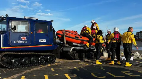 RNLI/Eve Kemp Seven volunteers in yellow jackets and boots surround the lifeboat - which is attached to the back of a blue vehicle - on a dry tarmac area near West Kirby beach