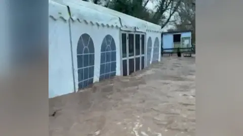 Otterton Mill A marquee sits with flood water rising high next to it, the flood water is a brown colour.