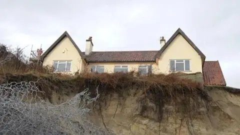 Jamie Niblock/BBC A view of The Chantry property taken from the beach below. Is is a large detached home with two gables at either end. There is a sandy cliff that has eroded and is now very close to the house. Rough grass grows over the edge of the cliff, but it is mostly sand. There is wire netting in the foreground.