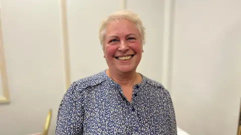 A woman in a patterned top and fair hair.