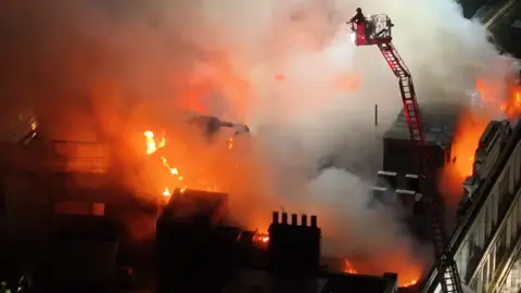 UKNIP A drone shot of someone on an aerial platform shooting water at flames on the roof of the building.