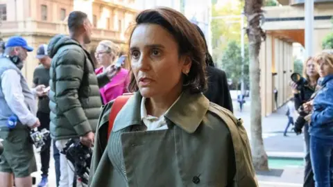 EPA A middle-aged woman with shoulder-length brown hair, wearing a khaki jacket with a crowd of photographers behind her
