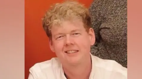 A man with blonde hair. He is looking at the camera and smiling. Behind him is an orange background.