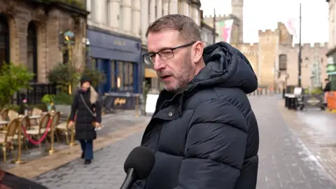 A man in a black puffer jacket and black glasses is speaking into a microphone on the street. Cardiff Castle and multiple bar/restaurants can be seen in the background.