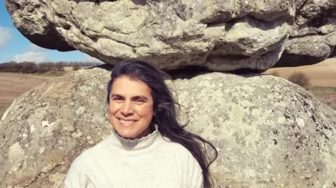 Deborah Maddison A close-up of Deborah in a field standing in front of a two large stones balancing on top of one another. She is smiling and her long, dark hair appears to be blowing in a gentle wind.