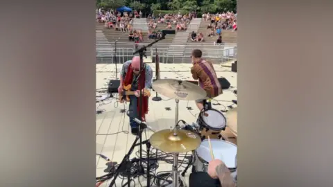 Telford and Wrekin Council A band perform on stage with the circular seating of the amphitheatre visible in the background. People are sitting some way away watching the band which includes a drummer, guitarist and singer. Cables and equipment can also be seen.