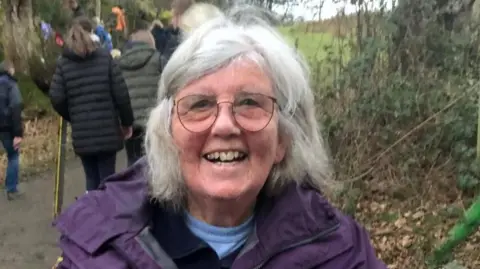 Maureen Rigg Maureen Rigg has neck length white hair, glasses, two blue tops and purple coat. She is smiling. She has people walking away from her behind her and undergrowth to the side of her.