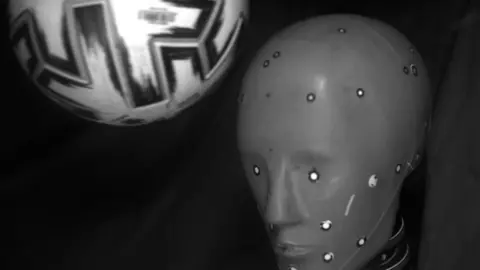 A black and white image with a grey test dummy head and a white and black football about to collide with it. The background appears to be a black curtain.