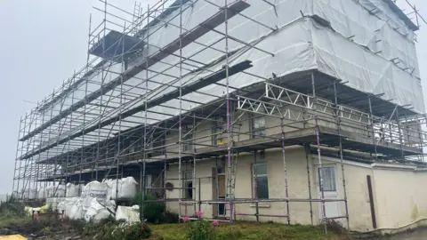 Three houses covered in white scaffolding and large sand bags. A small patch of grass can be seen in front of the houses with a misty sky in the background.