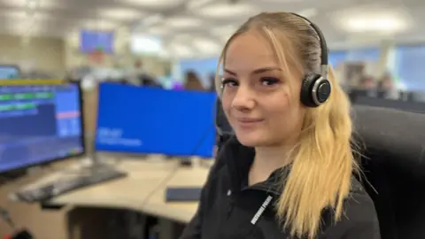 A young woman with long blond hair wearing a headset and Wiltshire Police lanyard and smiling to camera, sitting in front of a computer in a call centre with a blurred background