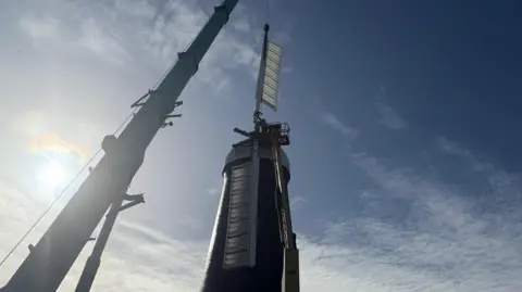 A crane is mid-way through attaching white sails back to a windmill. The windmill has a white cap and black side and the sun is shining on it.