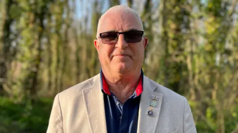 Joe Abbotts looking at the camera with trees behind him. He has short white hair, is wearing sunglasses, a cream blazer and a navy polo shirt.