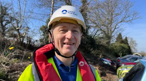 John is smiling at the camera and wearing a white hard hat, and red and high-vis safety gear. He is stood outside with trees behind him. The sun is shining.