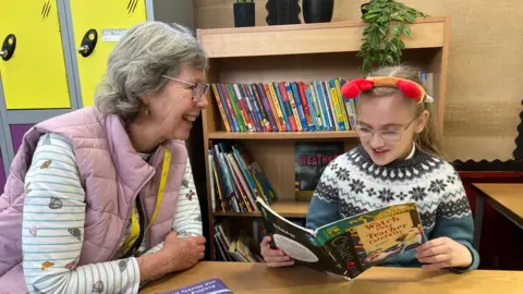 A classroom setting with a wooden table and bookshelves filled with colourful children's books in the background. A person wearing a pink quilted vest and striped top sits beside a child who is holding an open book titled 'Watch Your Teacher Carefully'. The child is wearing a festive headband with red antlers. On the table are reading materials, including a booklet and a sheet with coloured circles.