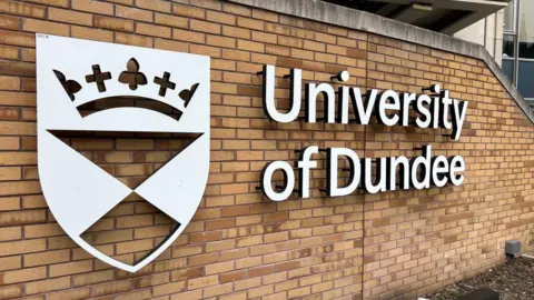 A white sign on a brick wall with a crest and the words University of Dundee
