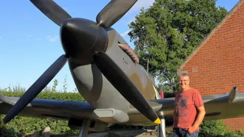 David Price David Price standing next to his spitfire. He has short white hair and glasses and is wearing a red t-shirt. Behind him is a red-brick gable end of a house. The spitfire is painted a brown-grey colour.